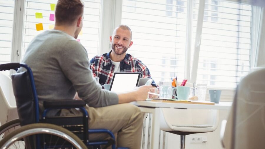 Mitarbeitender im Rollstuhl diskutiert mit einem Arbeitskollegen an einem Besprechungstisch. Auf dem Tisch aufgeklappte Laptops, Schreibutensilien, Tassen und Gläser.