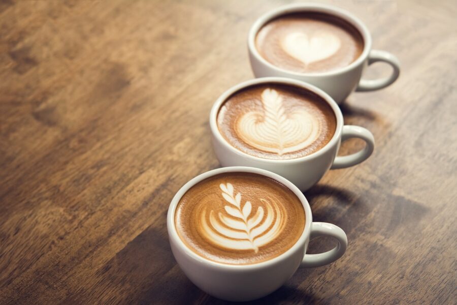 drei Tassen Kaffee mit Milchschaum stehen in einer Reihe hintereinander. Der Milchschaum ist in jeder Tasse unterschiedlich verziert.