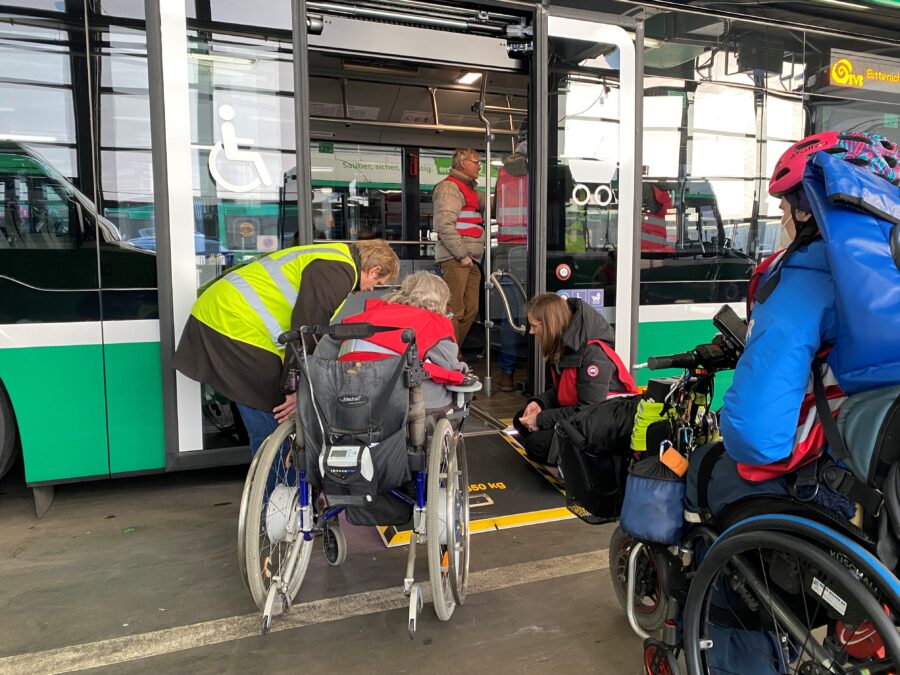 Zwei Rollstuhlfahrende betreten einen Bus. Ein BVB-Mitarbeiter hilft ihnen beim Einstieg.