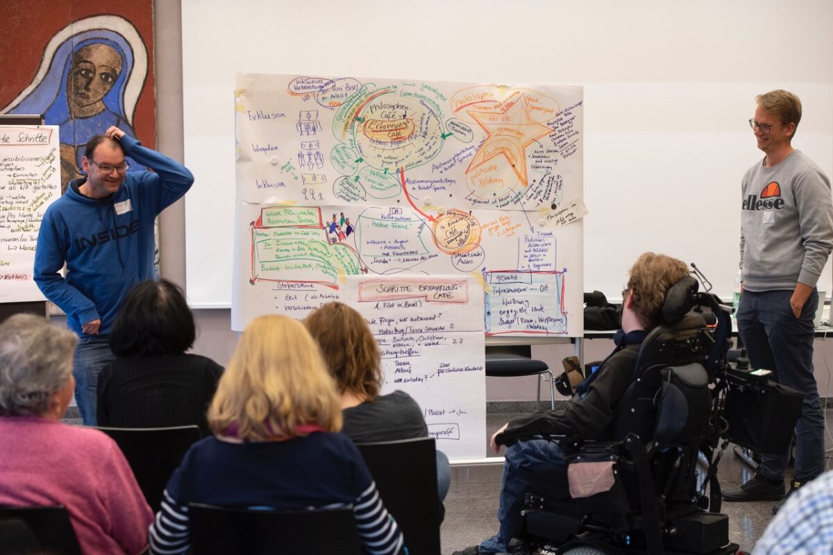 Verschiedene Menschen nehmen an einem Workshop teil. Ein farbiges Flipchart im Zentrum hält die Ergebnisse fest.