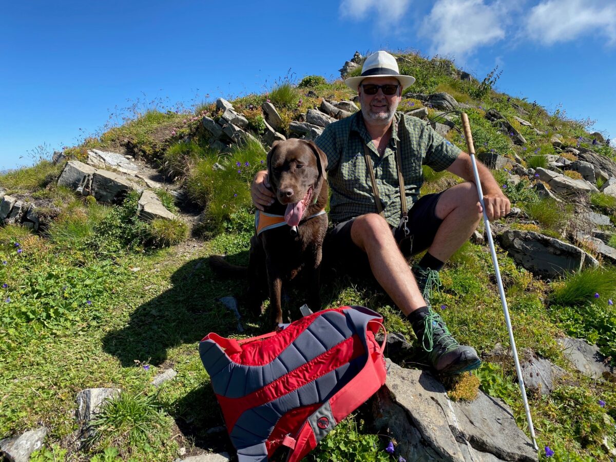 Das Bild zeigt einen blinden Mann, der gemeinsam mit seinem Blindenführhund während einer Wanderung in den Bergen eine Pause macht