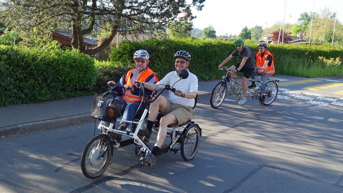 Im Vordergrund fahren zwei Männer auf einem Elektro-Dreirad. Im Hintergrund fahren zwei Männer auf einem Tandem-Velo.