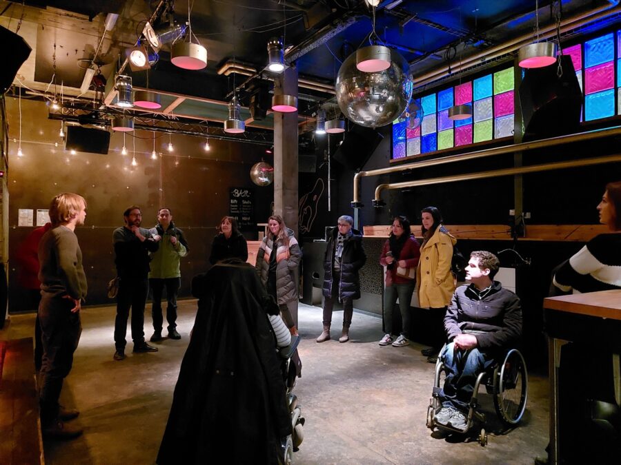 Eine Gruppe von 10 Personen, davon zwei im Rollstuhl, stehen im Kreis in einem Partyraum. Eine Person steht erhöht und erzählt den Teilnehmenden, die Teilnehmenden hören interessiert zu.