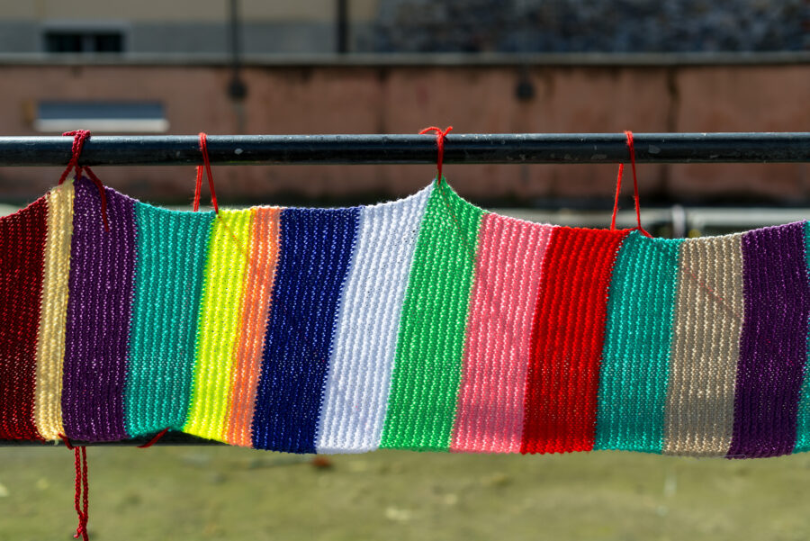 Ein Schal, der aus verschiedenfarbigen Wollen gestrickt wurde. Der Schal hängt an einem Geländer.