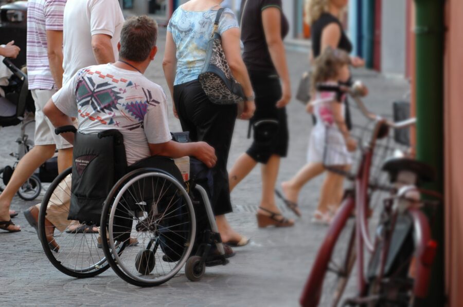 Ein Mann im Rollstuhl befindet sich im Vordergrund des Bildes. Er ist seitlich von hinten zu sehen. Er ist in mitten einer Stadt. Rundherum gehen andere Personen.