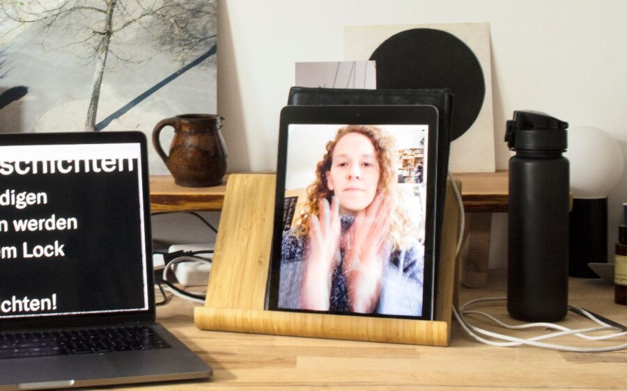 Auf einem Tablet ist das Bild einer Frau abgebildet. Das Tablet steht auf einem Tisch zwischen einem Laptop und einer Flasche.