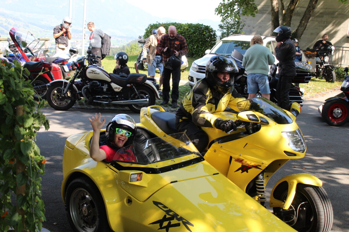 Auf dem Bild sind mehrere Motorräder mit Seitenwagen und Personen mit und ohne Motorradkleidung zu sehen. Im Vordergrund fährt ein gelbes Motorrad mit Seitenwagen gerade los. Der Fahrer trägt eine gelb-schwarze Motorradjacke und einen schwarzen Helm mit aufgeklapptem Visier. Im Seitenwagen sitzt eine lachende Person in rotem T-Shirt, einem schwarzen Helm und einer grünen Sonnenbrille. Sie hebt den rechten Arm zum Gruss.