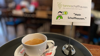 Auf dem Bild ist eine gefüllte Tasse Kaffee auf einem runden Tablett zu sehen. Daneben steht Bildhalter mit einer Karte mit dem Logo der tanneschaffhausen und dem Text «Mein Schaffhausen».