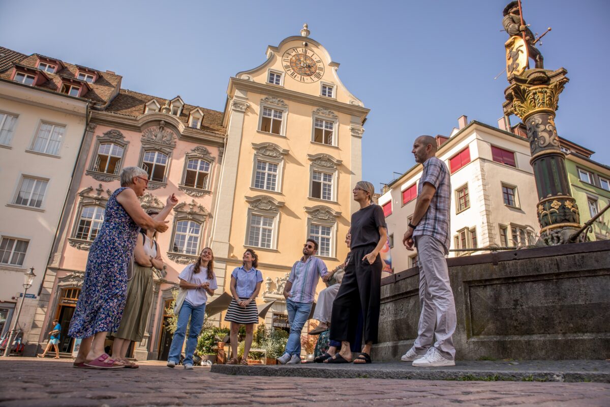 Bild vom Fronwagplatz mit Brunnen und Fronwagturm. Menschengruppe die von einer Reiseführerin Informationen erhalten.