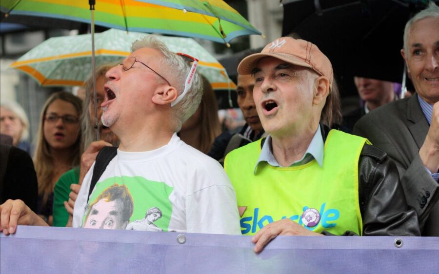 Auf dem Bild sind mehrere Personen zu sehen. Im Fokus stehen zwei ältere Männer. Sie halten zusammen ein Plakat. Es handelt sich um eine Demonstration.