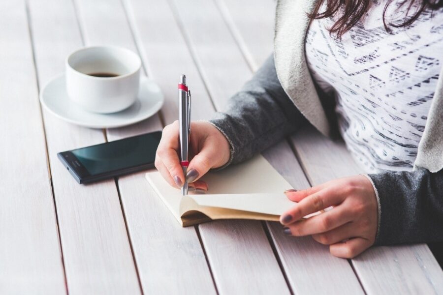 Eine Frau, die bei einer Tasse Kaffee in ein Notizbuch schreibt. Neben ihr liegt ein Handy.