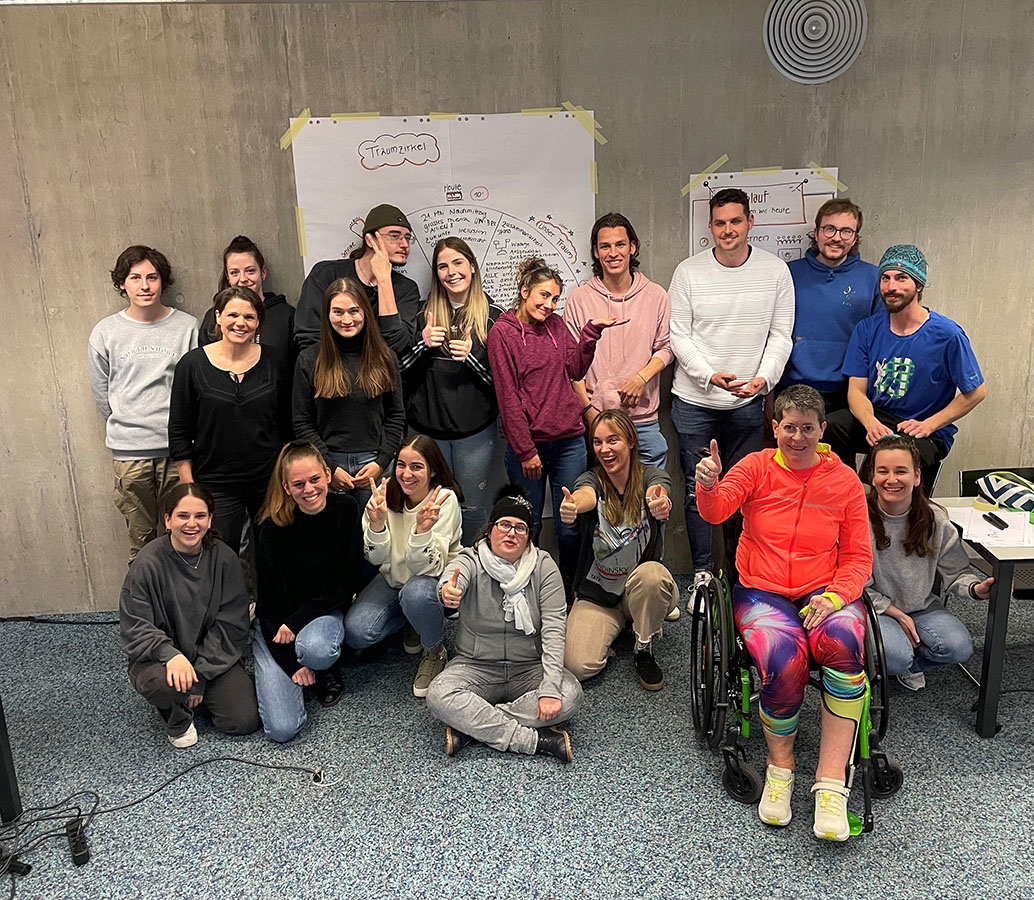 Studierende und Menschen mit Behinderungen stehen vor einer Wand in einem Raum der Hochschule für Soziale Arbeit in Siders. Im Hintergrund ist nicht lesbar das Plakat zu sehen, auf dem die Projektplanung beschrieben und in Kreisform gezeichnet ist.