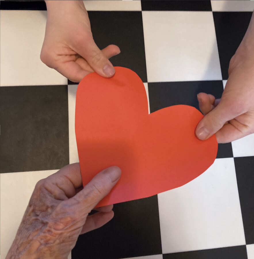 Drei Hände von Menschen unterschiedlichen Alters, die zusammen ein rotes Herz aus Papier halten. Der Hintergrund ist schwarzweiss-kariert.