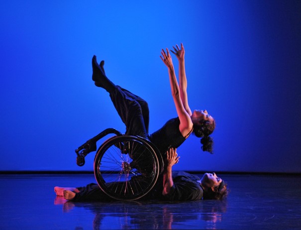 Zwei Personen auf einer Bühne. Eine Person liegt auf dem Rücken und stützt eine Person mit Rollstuhl, die sich nach hinten fallen lässt und die Arme und Füsse nach oben streckt.