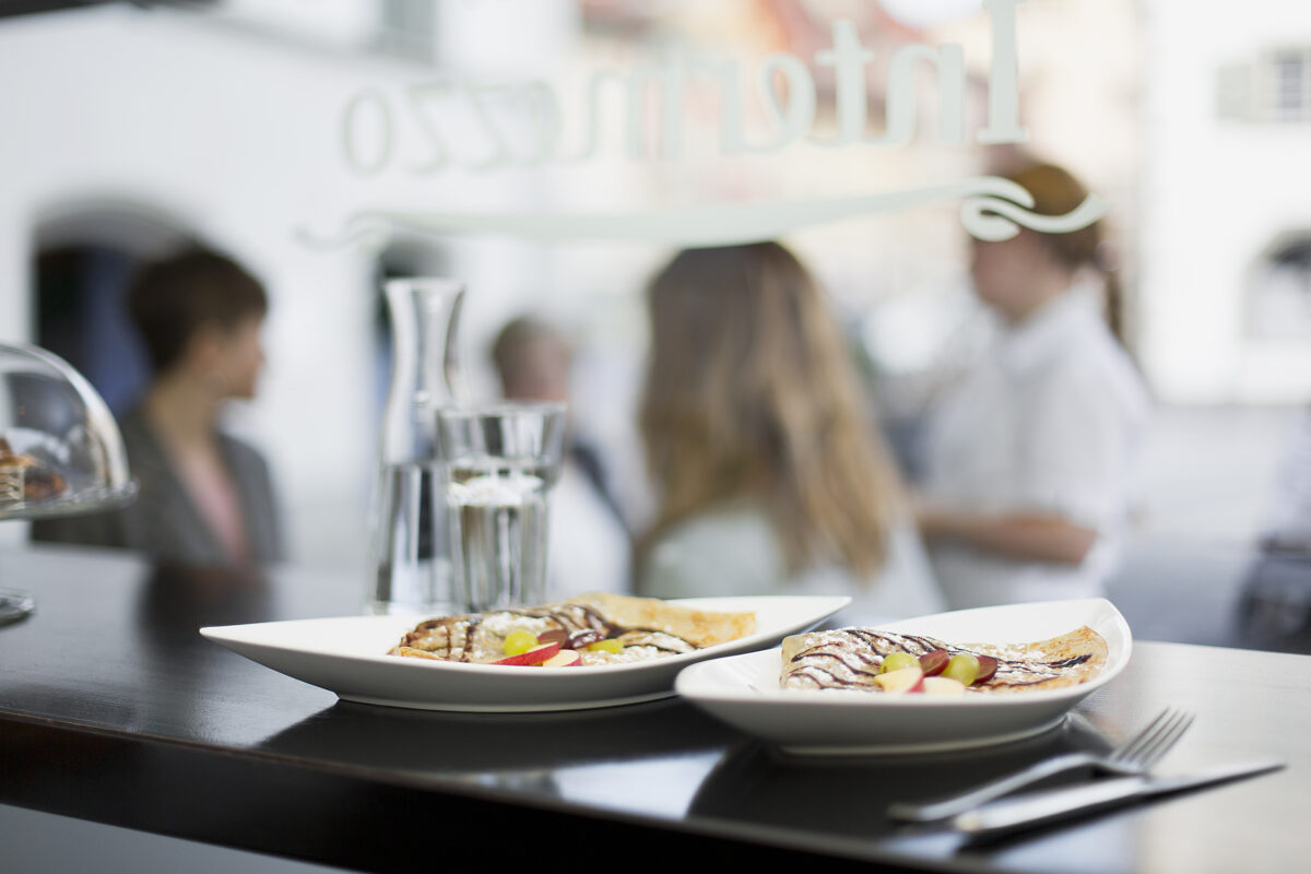 Zwei Teller mit Crêpes im Fokus, im Hintergrund sind Menschen zu erkennen.