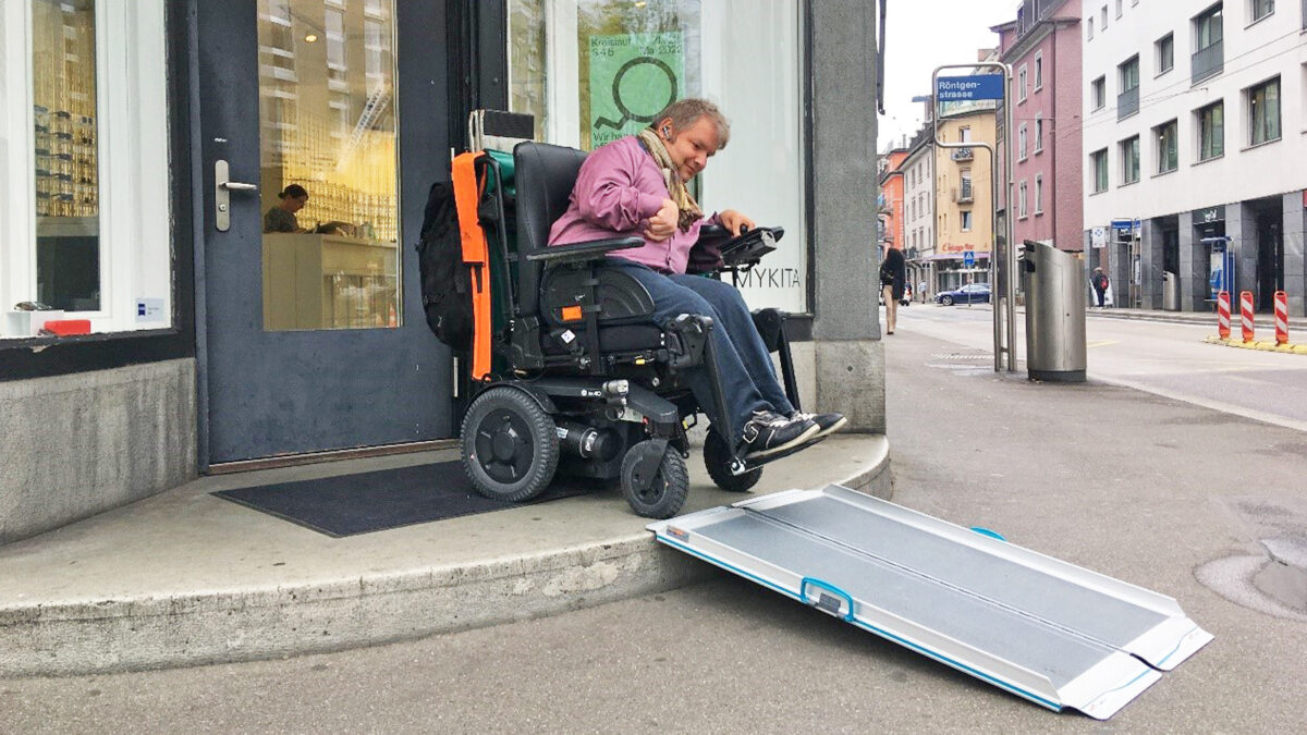 Mann im Elektrorollstuhl fährt über eine faltbare Rampe aus einem Geschäft. Der Eingang hat eine Stufe.