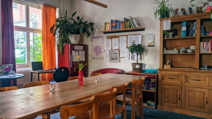 Ein einladender Raum empfängt die Besuchenden beim Eintreten in den Treffpunkt Nordliecht. In der Mitte ist ein langer hölzerner Tisch zu sehen, mit 7 Holzstühlen. Er bietet Platz für 8 Personen und lädt zum gemeinsamen Essen und Austauschen ein. Auf der rechten Seite steht ein hohes Buffet mit Tablaren, auf denen Bücher und Geschirr aufgereiht sind. Auf der linken Seite sieht man ein Fenster mit einem orangenen und violetten Vorhang, diese verleiden dem Raum eine gemütliche Atmosphäre. Eine Pinwand und ein gut bestücktes Bücherregal hängen an der Wand zwischen Buffet und Fenster. Der Raum ist mit Pflanzen ausgestattet, eine Blume steht in einer Vase auf dem Tisch.