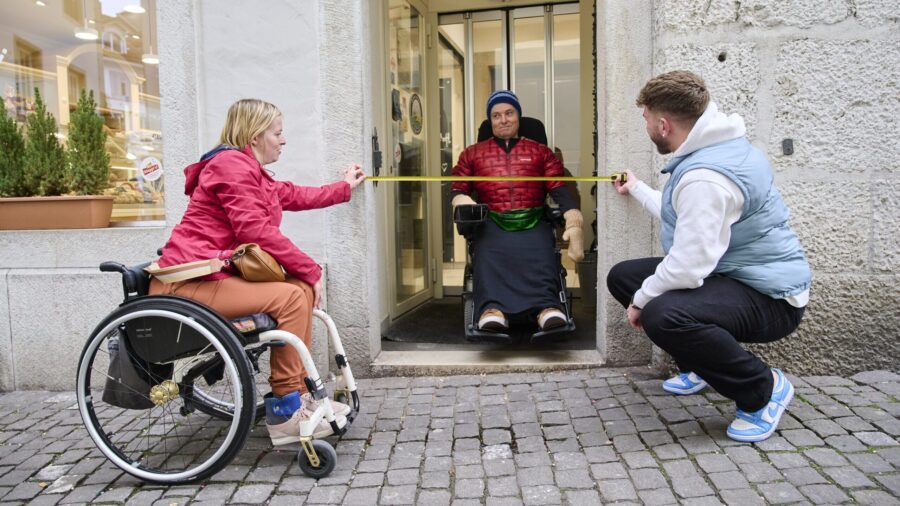 Zwei Personen messen mit Messband die Breite einer Haustüre. Mann steht mit Rollstuhl im Türrahmen..