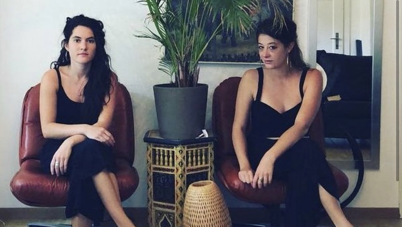Zwei Frauen sitzen im Wohnzimmer. Zwischen den beiden Frauen steht eine Palme.