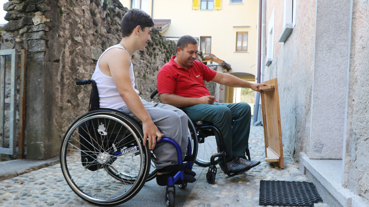 Zwei Menschen im Rollstuhl möchten mittels einer Rampe über eine Türschwelle gelangen.