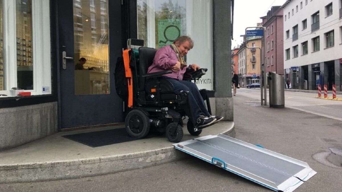 Ein Geschäft hat eine temporäre Rampe aufgebaut. Ein Mann im Rollstuhl fährt diese Rampe hinunter.