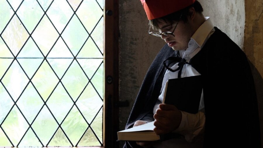 Person die vor einen Fenster steht und ein Buch liest. Sie trägt einen roten Hut und eine Brille.