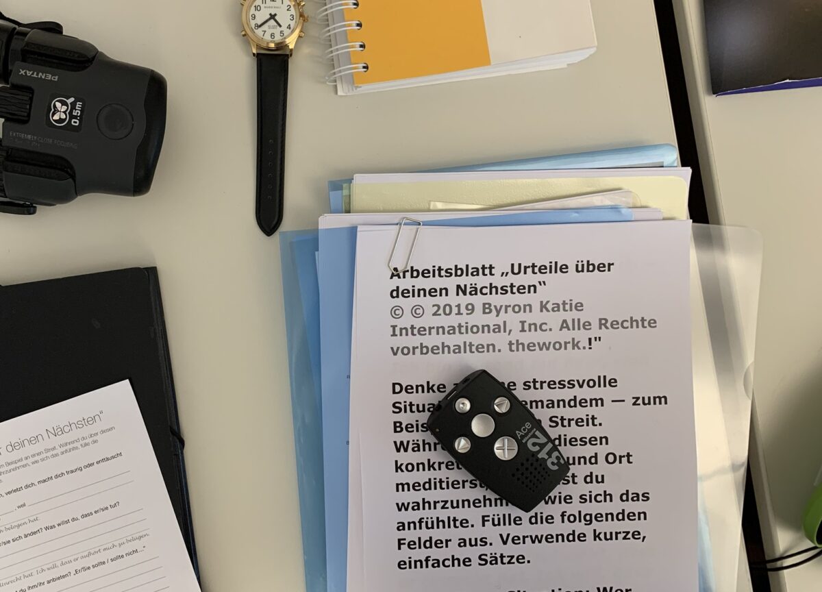 Auf einem Tisch liegt ein Stapel von Blättern, die in grosser Schrift bedruckt sind. Auf dem Stapel liegt ein kleines elektronisches Gerät mit fünf Tasten. Neben dem Stapel liegen zudem eine Uhr, weitere Blätter, ein gelbes Ringheft und ein weiteres schwarzes, elektronisches Gerät.