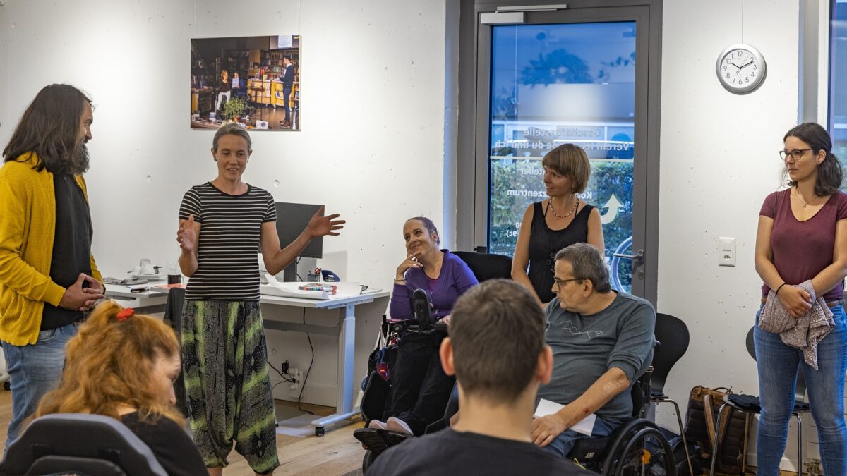 Foto einer Schulung: Eine Gruppe von Menschen im Kreis. Männer und Frauen, manche sind im Rollstuhl, manche stehen. Eine Frau erzählt etwas, die anderen hören lächelnd zu.