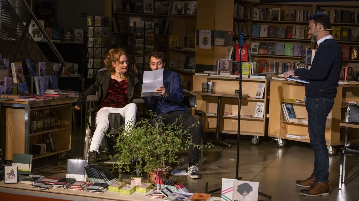 Man sieht die Autorin auf der Bühne, sie ist im Rollstuhl. Daneben sitzt ihre Dolmetsch-Assistenz und hält ein Blatt Papier in der Hand. Rechts ist der Moderator Nik Hartmann zu sehen, der das Buch der Autorin in den Händen hält. Im Hintergrund sieht man einen Bücherladen mit vielen Büchern.