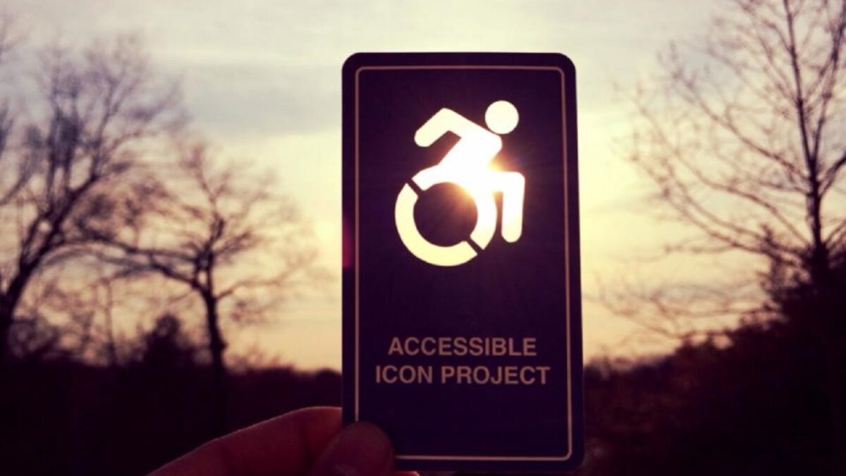 Auf der Fotografie hält jemand eine grosse Karte in die tief stehende Sonne. In die Karte mit der Beschriftung «Accessible Icon Projekt» ist ein Rollstuhl-Piktogramm gestanzt. Die Sonne strahlt dadurch durch diese Umrisse hindurch. Es handelt sich nichtum das gewöhnliche, statische Rollstuhl-Symbol. Hier ist die Person in Bewegung und der Rollstuhl wirkt rollend. Im Hintergrund sind unscharf mehrere grosse Bäume ohne Blätter zu sehen.