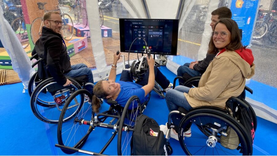 Eine Besucherin testet den Handbike Simulator und erhält von Menschen im Rollstuhl Tipps.