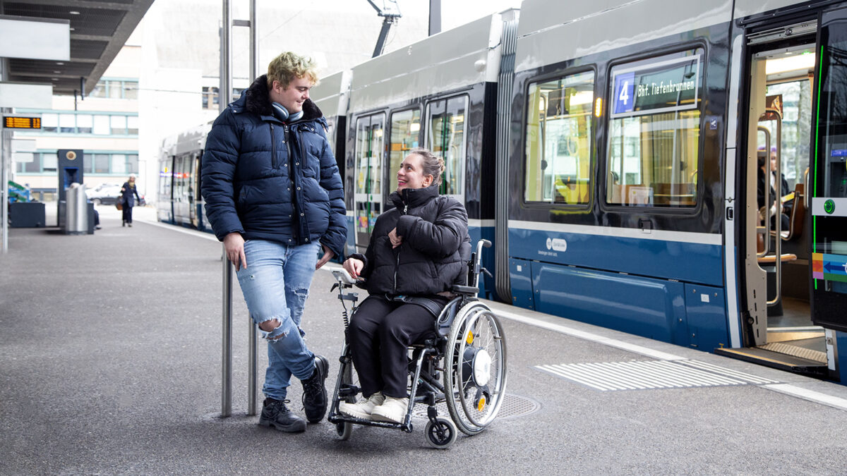 Ein stehender Mann und eine sitzende Frau im Rollstuhl sind vor einem Tram. Sie schauen sich an und lächeln.