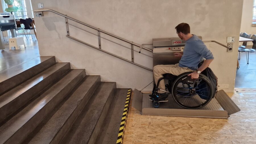 Ein Mann sitzt im Rollstuhl. Er befindet sich vor einer Treppe, auf dem Rollstuhl-Lift und testet diesen.