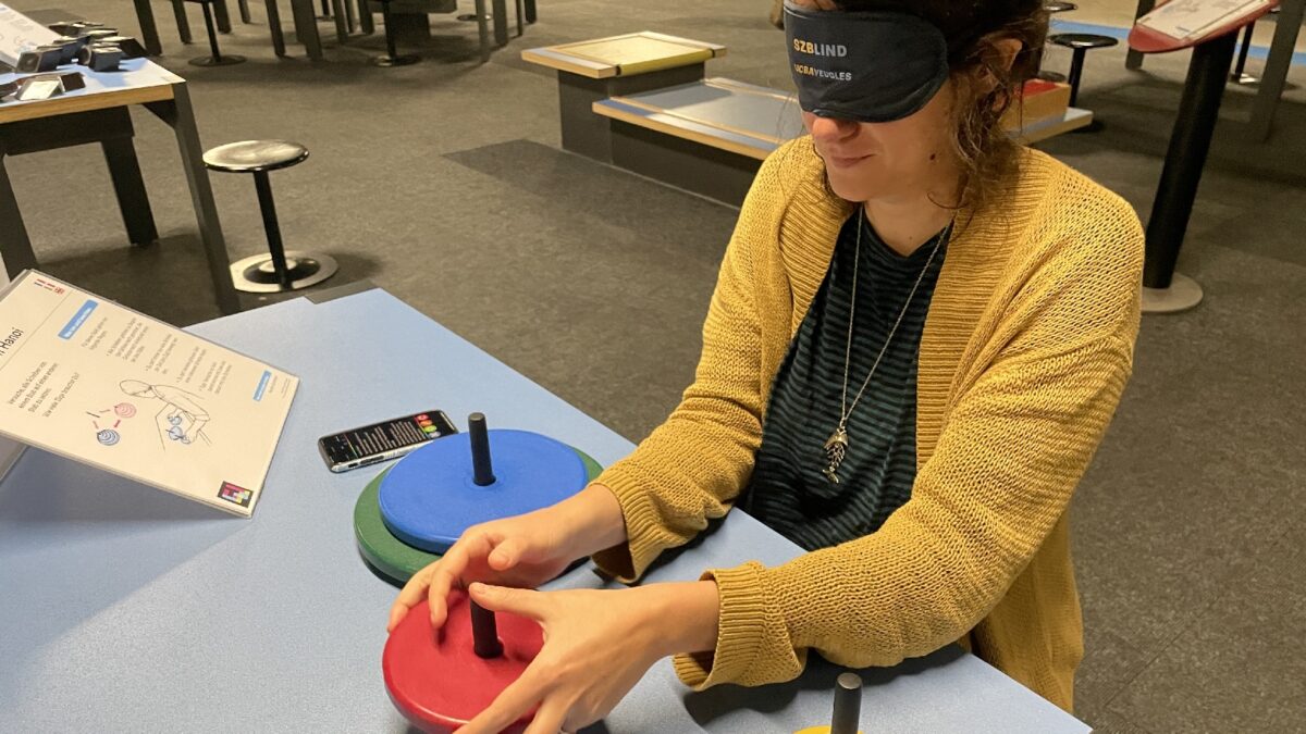 Eine Person sitzt an einem Tisch in der Ausstellung. Sie trägt eine schwarze Augenbinde mit der Aufschrift SZBLIND. Auf dem Tisch sind drei Stäbe befestigt, auf den Stäben hat es verschiedenfarbige Scheiben (blau, grün, gelb, rot und weiss). Die Person nimmt die rote Scheibe von einem Stab. Neben der Person liegt ein Handy, davor steht eine Tafel mit der Anleitung und einem farbigen NaviLens-Code.