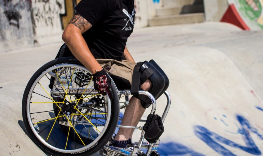 Auf dem Bild ist eine Person im Rollstuhl zu erkennen. Die Person befindet sich in einem Skatepark. Sie trägt Handschuhe und Knieschoner.