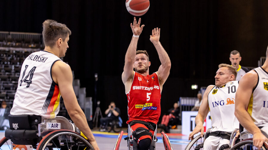 Wurfaktion im Rollstuhlbasketball-Spiel zwischen der Schweiz und Deutschland. In der Mitte des Bildes befindet sich ein Mann im Rollstuhl. Er trägt ein rotes T-Shirt. Er streckt beide Arme über den Kopf und wirft den Basketball. Um ihn herum hat es weitere drei Männer im Rollstuhl. Sie alle befinden sich in einer Turnhalle. |