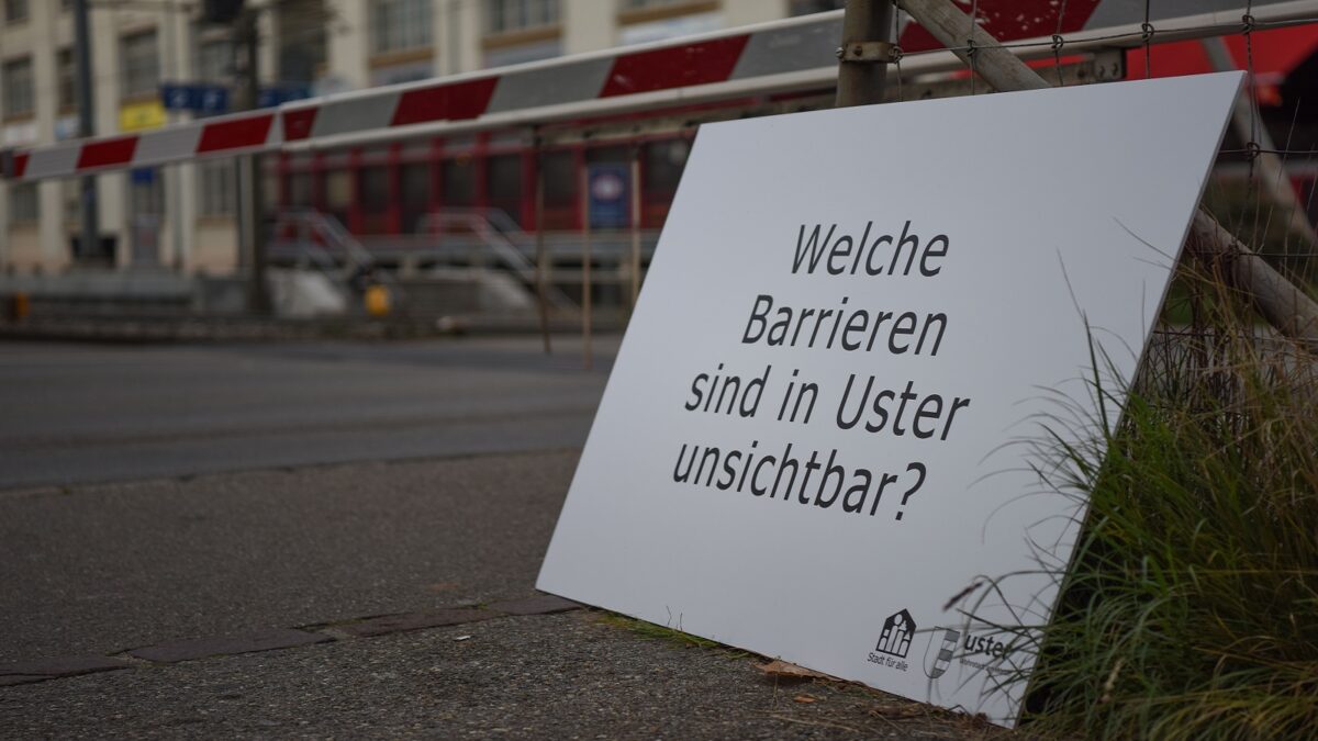 Auf dem Bild liegt ein weisses Schild angelehnt am Boden. Mit schwarzer Schrift steht: Welche Barrieren sind in Uster unsichtbar?