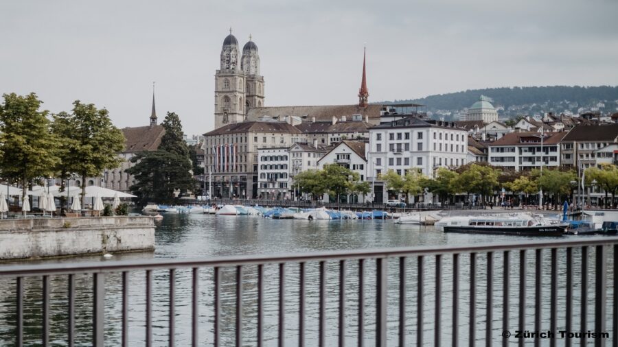 Wir befinden uns neben dem Hauptbahnhof Zürich. Das Bild wurde auf einer Brücke aufgenommen. Im Vordergrund kann man Wasser sehen, auf welchem zahlreiche Boote schwimmen. Am Ufer auf der linken Seite sind zwei Bäume und offene sowie geschlossene weisse Sonnenschirme. Im Hintergrund sieht man die Stadt Zürich.