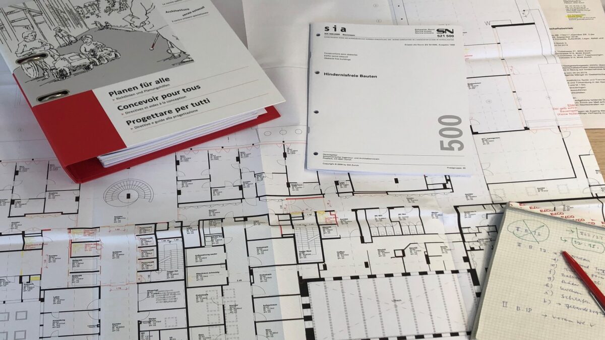 Das Bild zeigt einen Bauplan. Im oberen Bereich des Bildes liegen die Norm zum "Hindernisfreien Bauen" und ein Ordner. In der unteren rechten Ecke liegt ein Notizblock mit einem roten Stift.