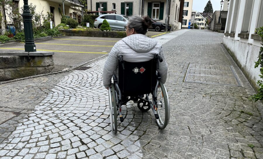 Eine Person sitzt im Rollstuhl. Der Rollstuhl steht auf einem Kopfsteinpflasterweg.