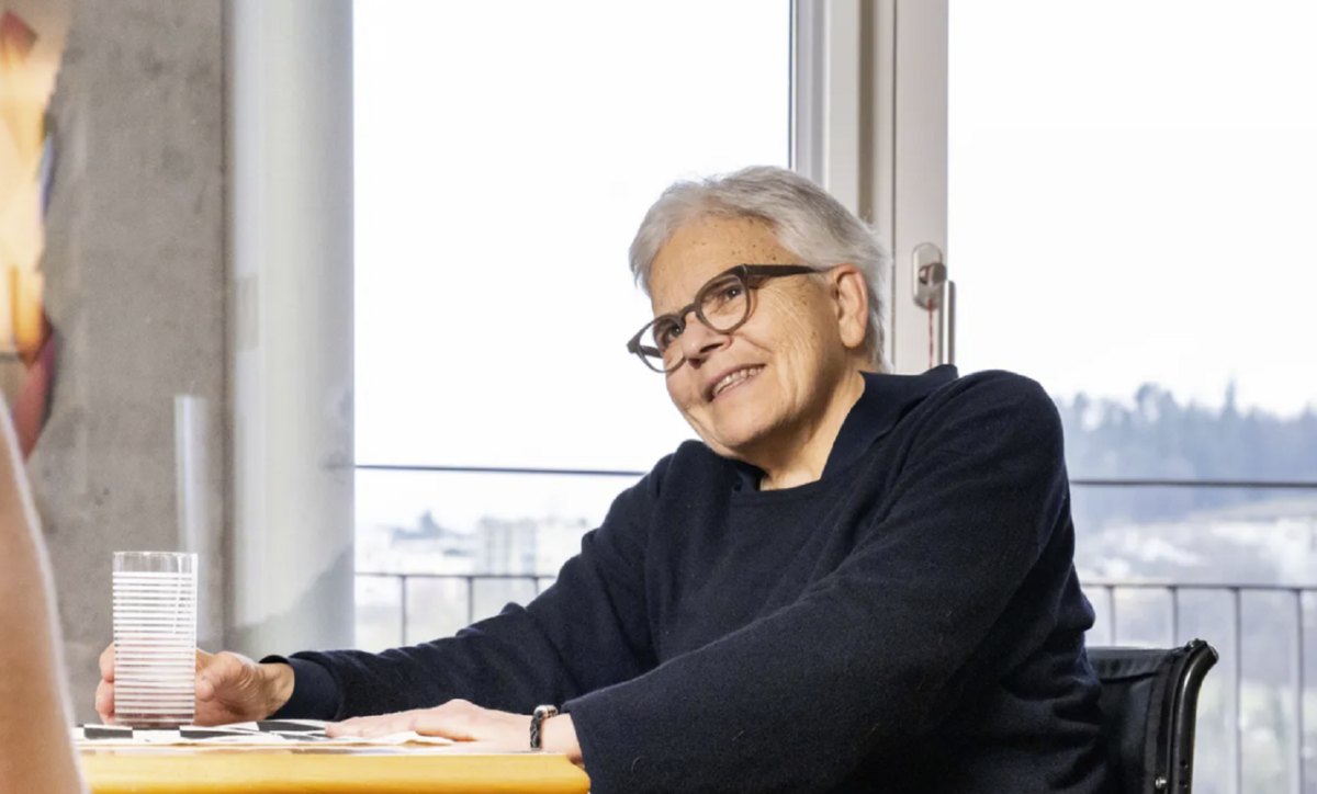 Eine Frau sitz an einem Tisch. Sie trägt kurzes graues Haar und eine schwarze Brille. In ihrer rechten Hand hält sie ein Glas.