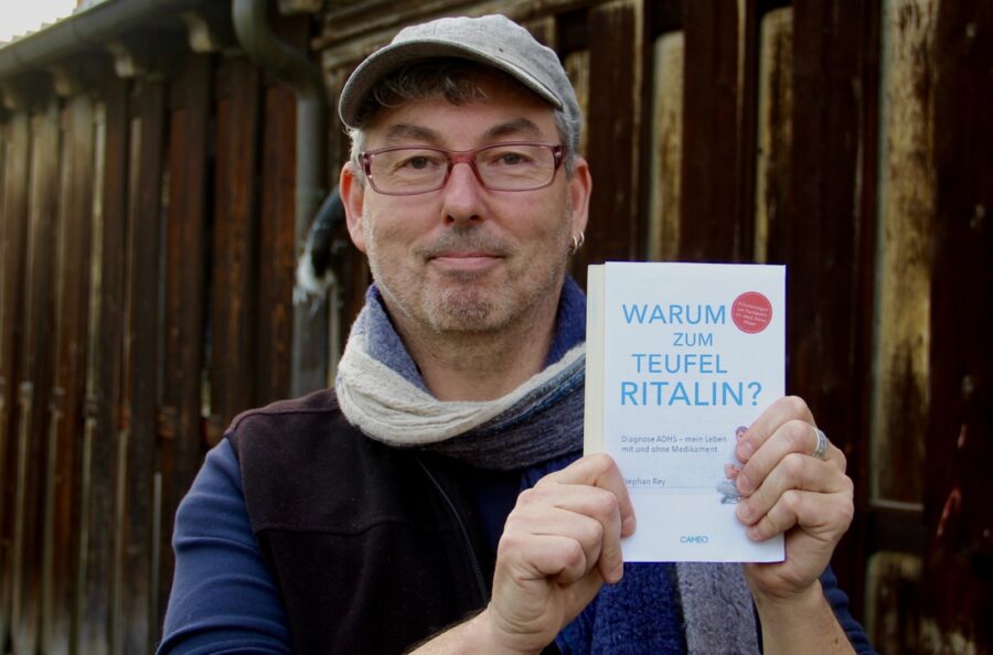 Portrait des Autor Stephan Rey. Er trägt eine graue Kappe und eine rote Brille. Sein Haar ist grau meliert. Um den Hals hat er ein blaues Halstuch. In den Händen hält er sein Buch «Warum zum Teufel Ritalin?»