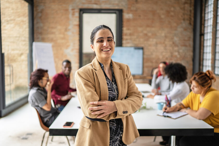 Eine Frau steht lächelnd in einem Sitzungsraum. Im Hintergrund sitzen mehrere Personen an einem Tisch und arbeiten gemeinsam.