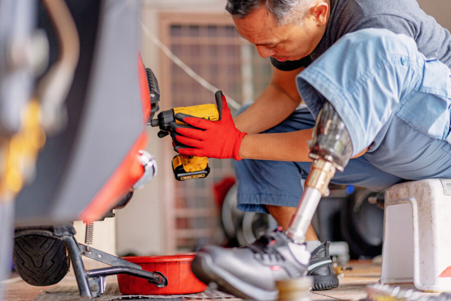 Ein Mann mit Beinprothese sitzt am Boden und arbeitet mit einem Akkuschrauber an einem Fahrzeug oder Gerät. Neben ihm stehen Werkzeuge und Material.