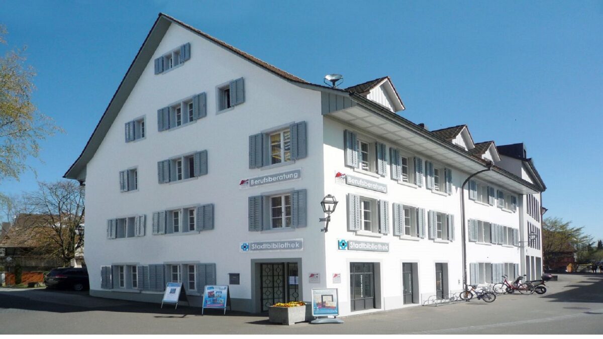 Auf dem Bild sieht man die Aussenansicht der Stadtbibliothek Bülach inkl. Schild «Stadtbibliothek», das an der Hausfassade hängt, sowie mehrere im Veloständer parkierte Velos auf der rechten Seite vor dem Gebäude.