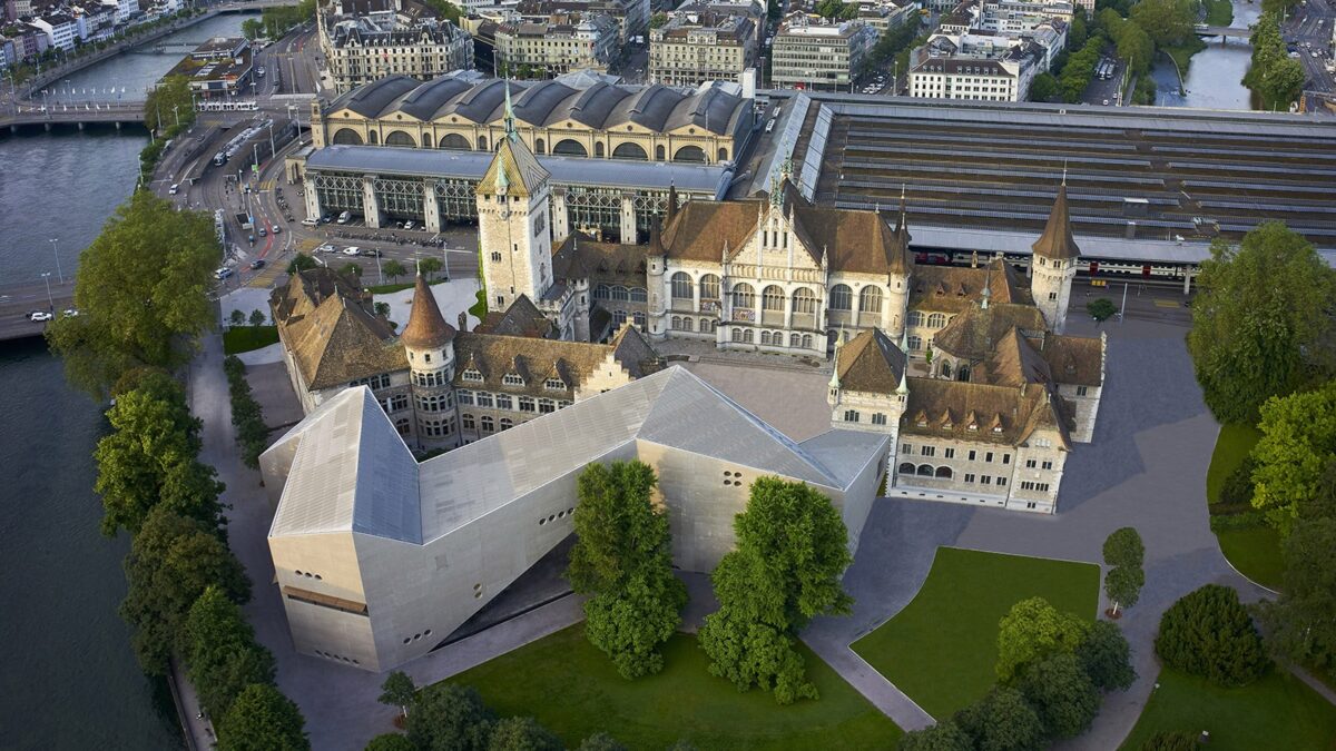 Landesmuseum und Hauptbahnhof aus der Vogelperspektive. Das Landesmuseum ist ein grosses Gebäude, welches aus einem älteren Teil von 1898 und einem angefügten Neubau von 2016 besteht. Zum schlossähnlichen Altbau gehören verschiedene Türme, die vorherrschenden Farben sind beige und braun. Der Neubau stellt mit seinen starken Linien und geraden Flächen einen Kontrast zum älteren Bau dar