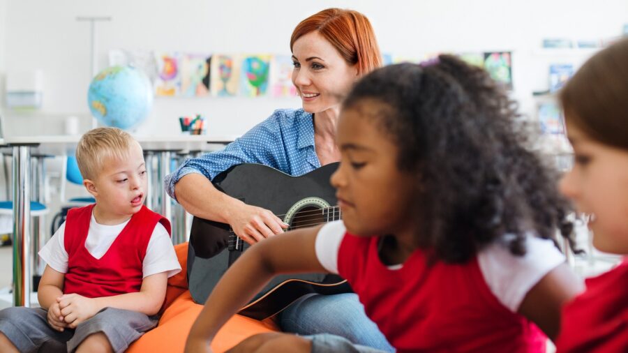 Eine rothaarige, freundlich lächelnde Frau sitzt mit drei Kindern in einem Schulzimmer.Sie spielt Gitarre. Der Junge rechts neben ihr hat Trisomie 21 und schaut auf ihre Finger. Ein dunkelhäutiges und ein hellhäutiges Mädchen, welche verschwommen abgebildet sind, sitzen links von der Frau. Alle scheinen der Musik zuzuhören