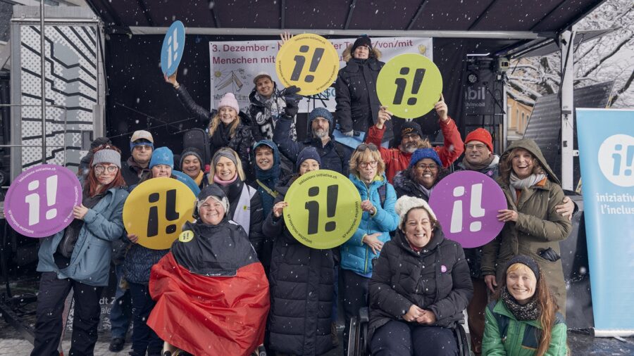 Eine Gruppe von Menschen mit und ohne sichtbare Behinderung halten bunte, runde Schilder mit dem Logo der Inklusions-Initiative. Alle lachen und tragen warme Winterkleider.