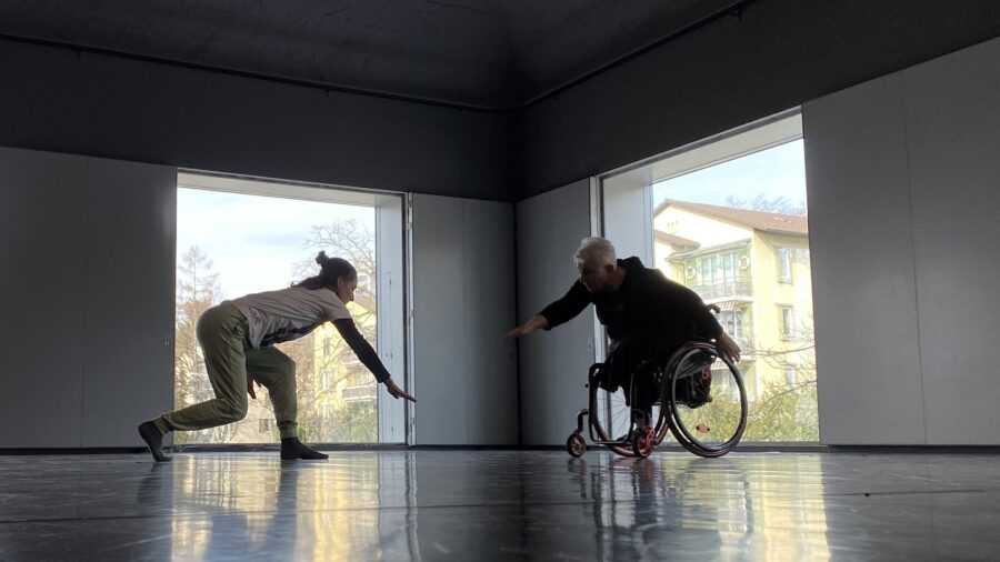 zwei Frauen, eine davon im Rollstuhl, tanzen in einem grossen Raum mit zwei grossen Fenstern