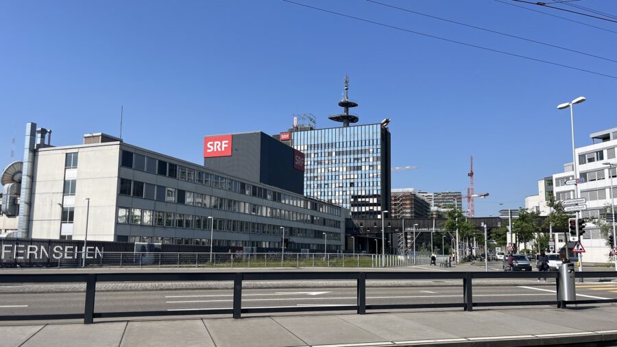 Auf dem Bild ist das Gebäude der SRG in Zürich abgebildet. Es ist ein grosses gläsernes Hochhaus, davor ein kleinerer schwarzer Gebäudeblock und im Vordergrund ein längliches graues Gebäude.